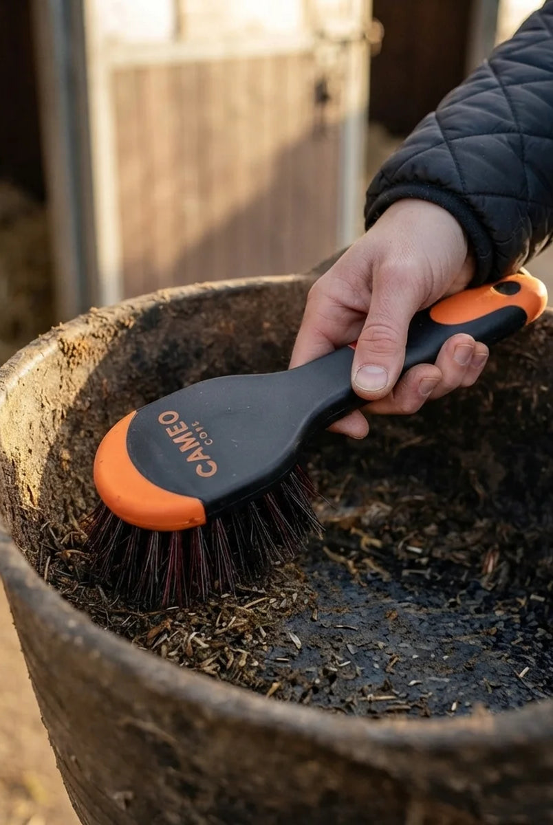 A lifestyle image featuring a person wearing a black quilted jacket brushing the inside of a rubber feed bucket with the orange-and-black CAMEO CORE brush.