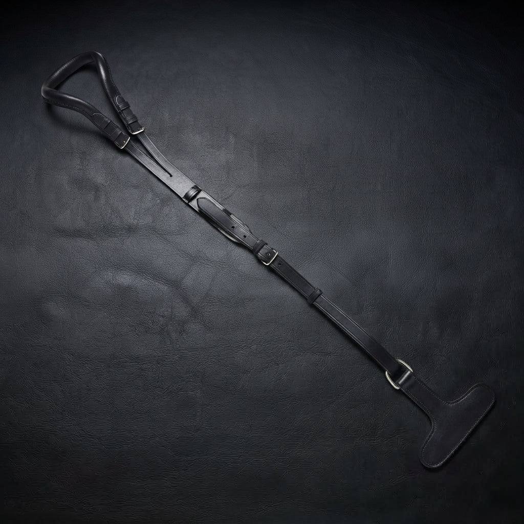 A black leather horse crupper with crupper T strap laid diagonally on a dark textured leather surface, featuring silver-toned metal buckles and a padded black handle.