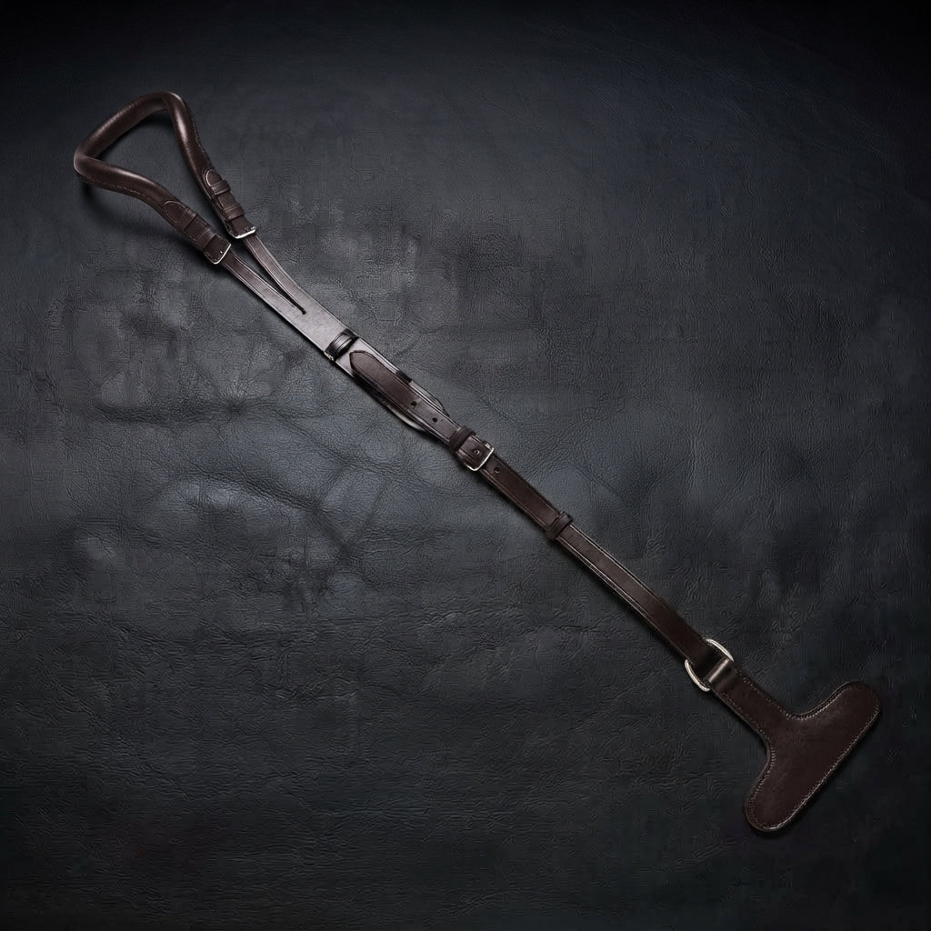 A dark brown leather crupper with T with metal buckles, laid diagonally on a dark textured background.
