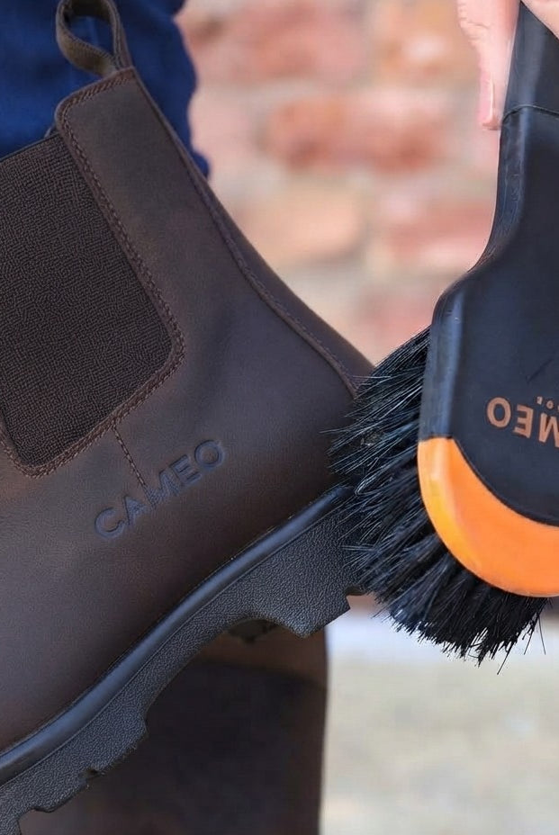 A detailed product photo showing a clean brown leather Chelsea-style yard boot being brushed with a black and orange CAMEO-branded brush. The boot features a subtle embossed CAMEO logo on the side and is paired with a realistic leg wearing navy blue equestrian riding tights.