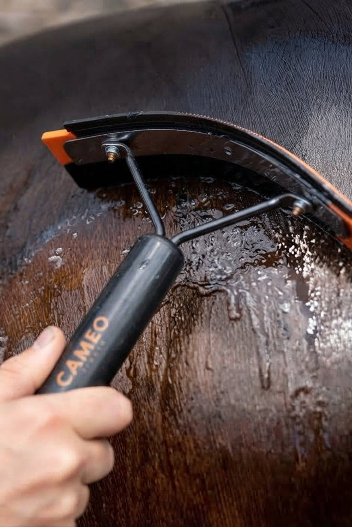 A close-up of a horse being washed with water using a black and orange horse grooming brush with a black handle. A human hand is holding the handle, which has the brand name 'CAMEO' printed on it.