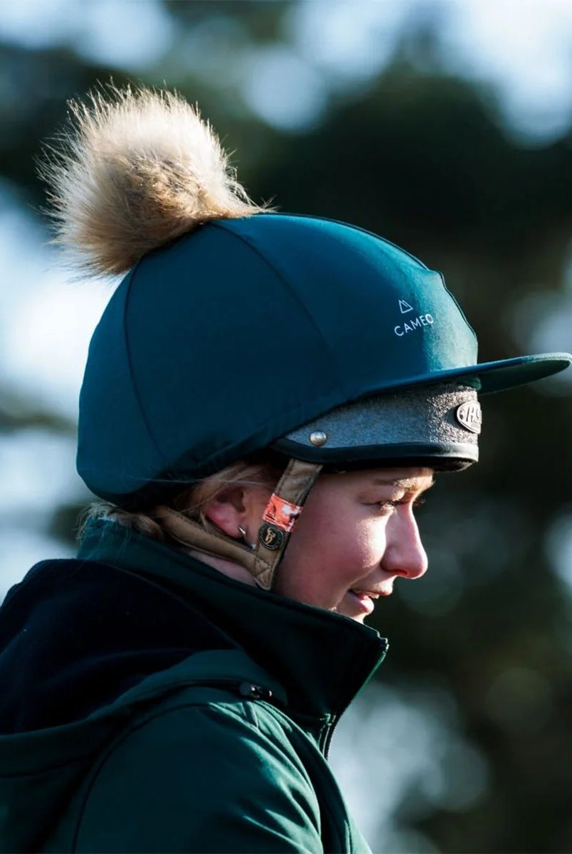 CAMEO Core Hat Silk in EverGreen worn by a rider featuring a detachable luxe faux-fur pom-pom and 4-way stretch CameoTech fabric for a wrinkle-free fit over a riding helmet. This stylish equestrian headwear includes a flexible peak and subtle CAMEO branding, perfect for matching with the EverGreen riderwear collection.