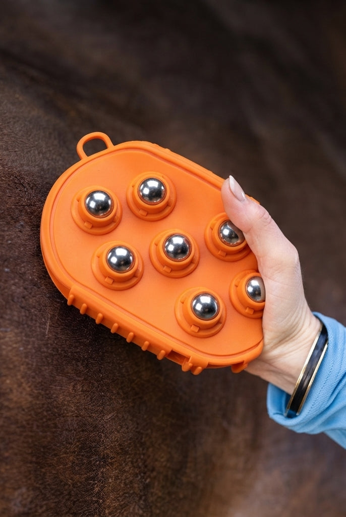 An orange horse grooming brush with six metallic ball nubs being held by a person's hand against a dark brown horse's coat.
