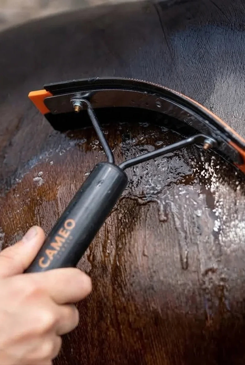 A close-up of a horse being washed with water using a black and orange horse grooming brush with a black handle. A human hand is holding the handle, which has the brand name 'CAMEO' printed on it.