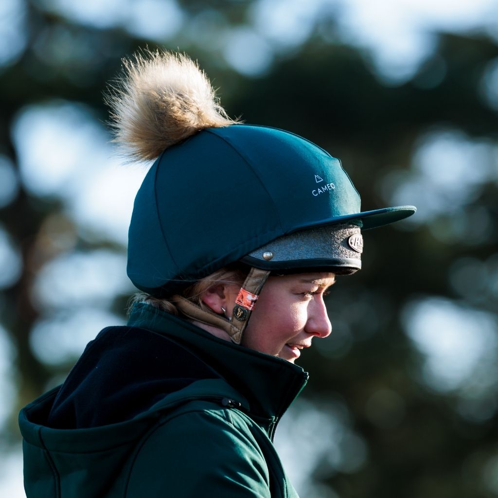 Profile view of an equestrian wearing a CAMEO EverGreen C417 hat silk featuring the white CAMEO logo and a fluffy faux-fur pom-pom.