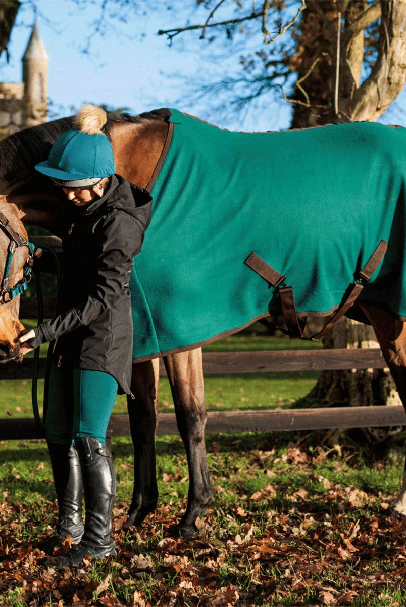 A woman in equestrian attire stands beside a brown horse outdoors, holding the horse's reins while wearing a teal horse blanket with dark brown leather straps. The setting is a grassy field with fallen leaves, wooden fencing, and trees under a blue sky.