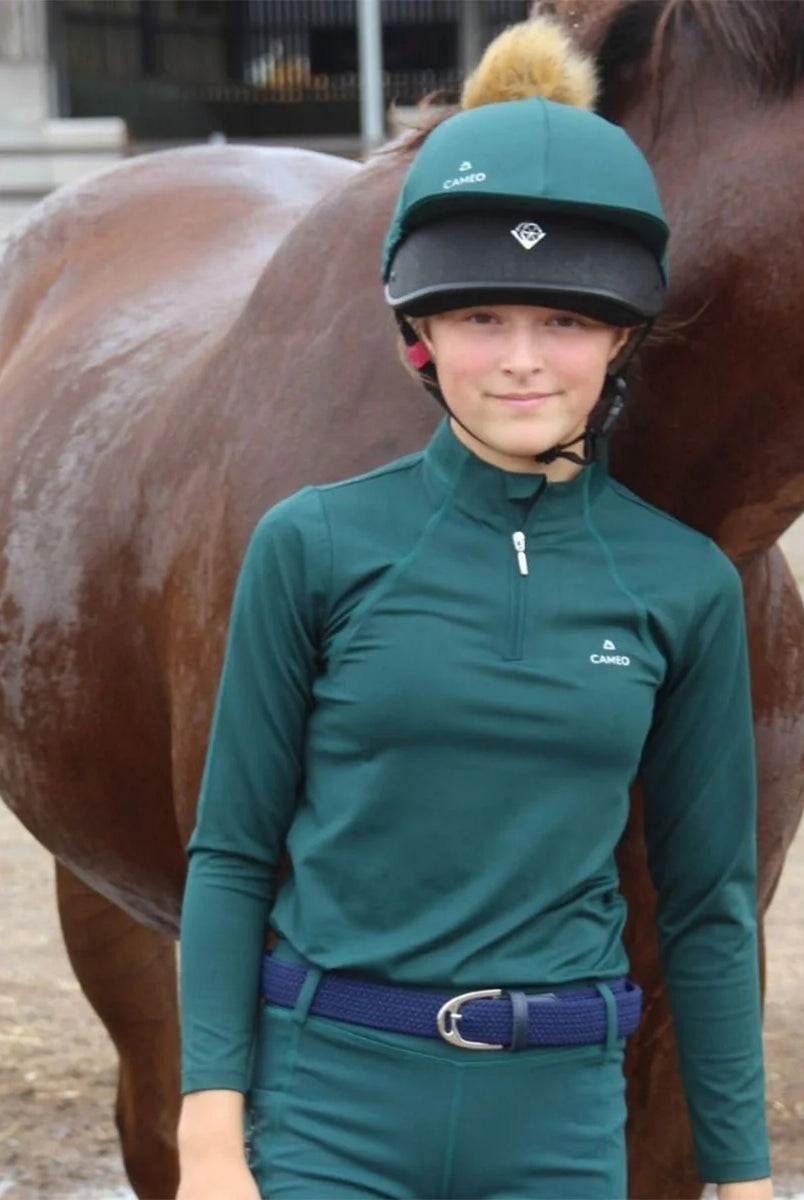 CAMEO Junior Core Baselayer in EverGreen featuring CameoTech 4-way stretch fabric and a quarter-zip stand collar. Worn by a young rider with a matching EverGreen hat silk, this moisture-wicking equestrian top offers active thermoregulation and flat-lock seams for training and competition in the UK.