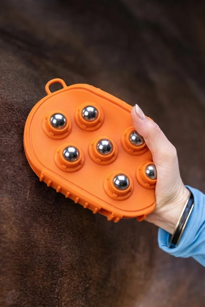 An orange horse grooming brush with six shiny silver metal balls, being held by a person's hand against a dark brown horse's coat.
