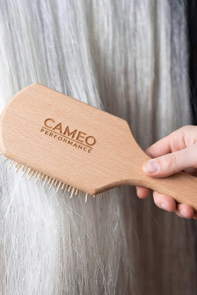 A hand holding a light natural wood paddle hairbrush engraved with 'CAMEO PERFORMANCE' against a background of white and black horse tail hair.