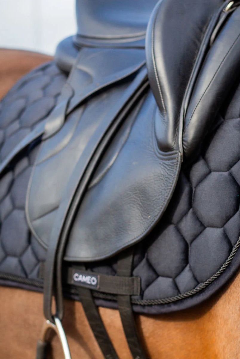 CAMEO Suede Dressage Saddlecloth in Black viewed in an angled close-up beneath a dressage saddle on a horse, highlighting the straight flap cut, honeycomb quilting, rope piping, and reinforced girth loop engineered for shock absorption and non-slip stability during intensive dressage schooling.
