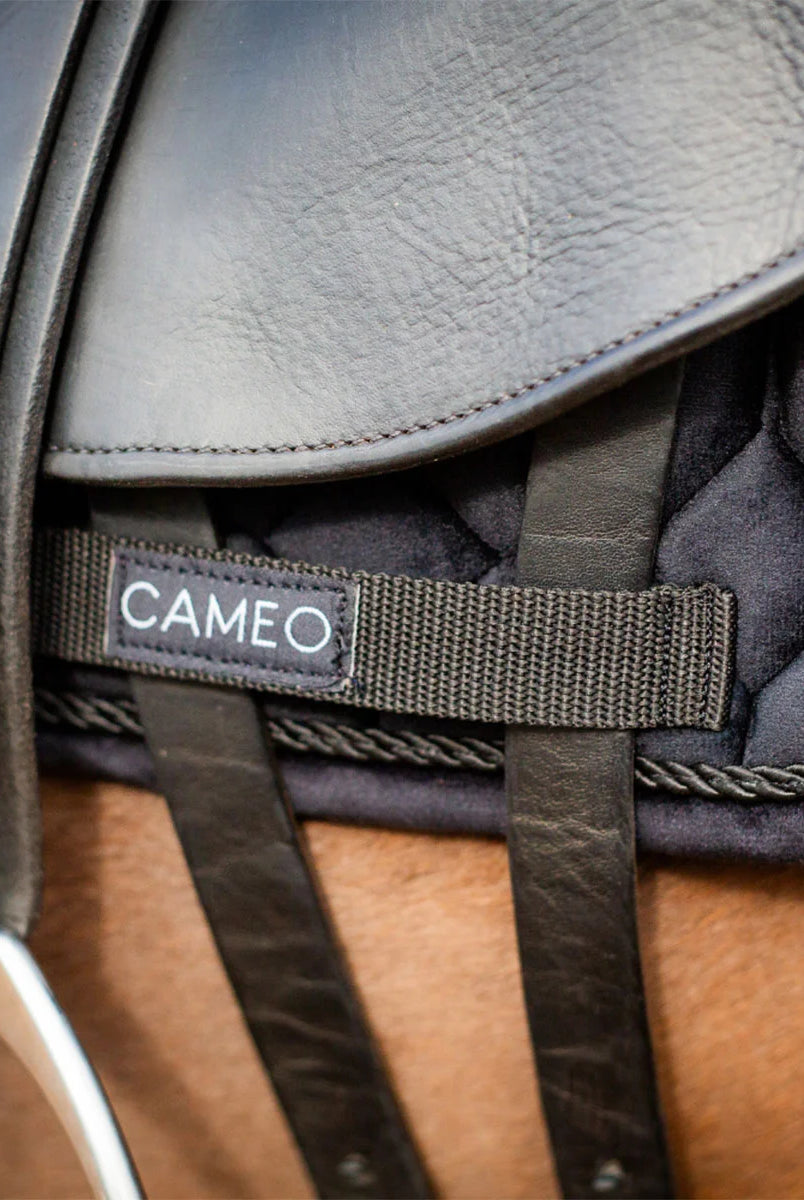 CAMEO Suede Dressage Saddlecloth in Black in a macro close-up beneath a saddle flap, showcasing the heavy-duty webbing girth protection loop with woven brand badge, leather billet straps, and rope piping engineered to prevent saddle friction wear and ensure non-slip stability during dressage riding.