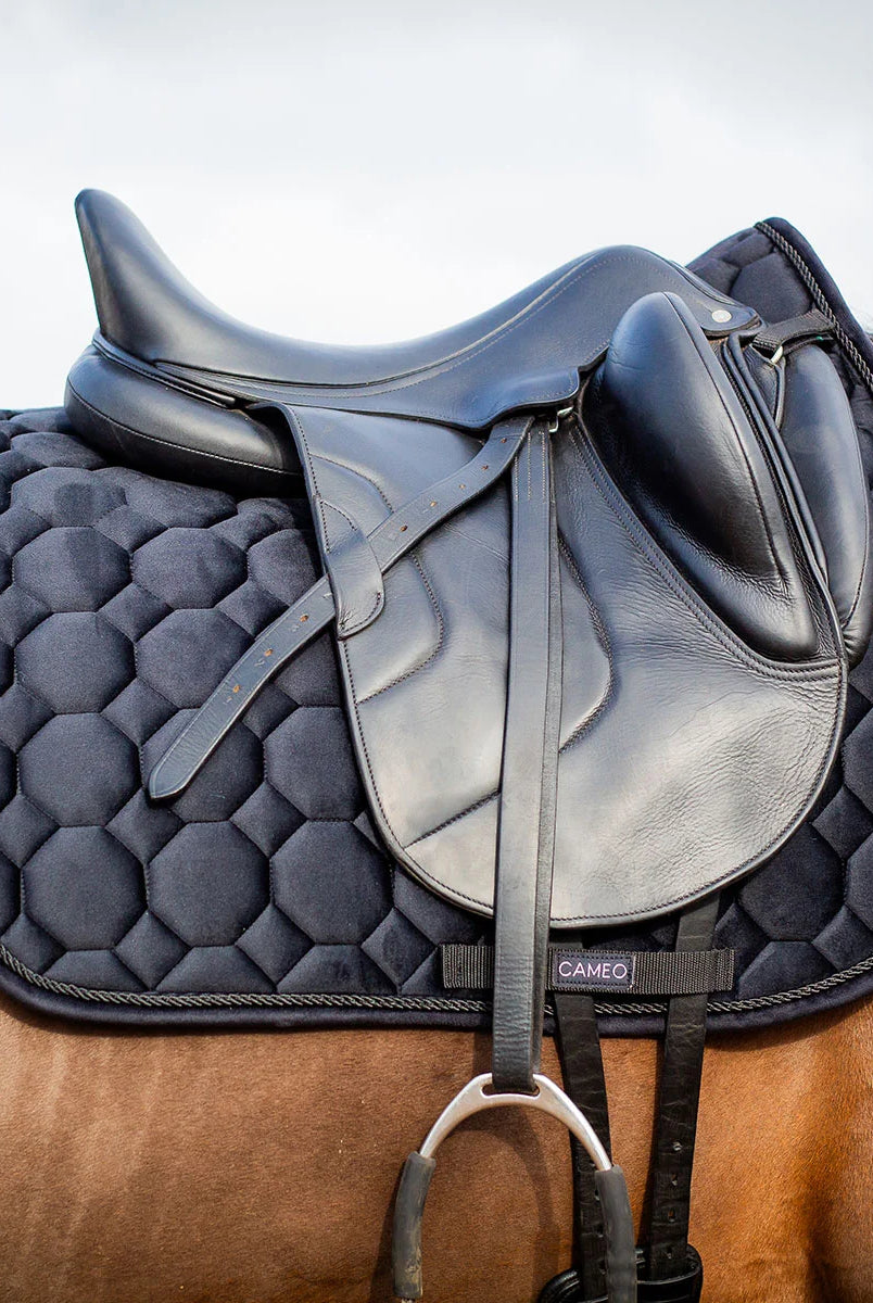 CAMEO Suede Dressage Saddlecloth in Black fitted beneath a black leather dressage saddle on a bay horse, showcasing the straight-cut square profile, hexagonal honeycomb quilting, rope piping, and branded girth protection panel engineered for shock absorption, spinal relief, and arena performance.