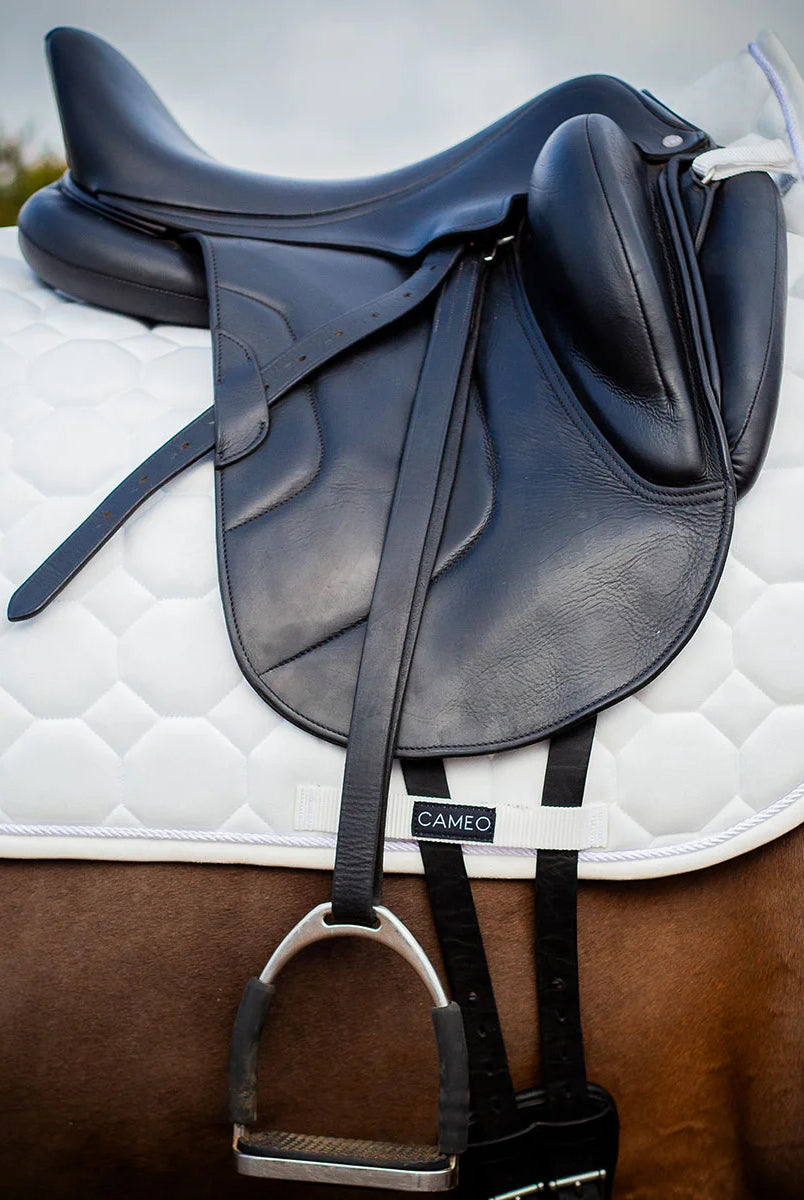 CAMEO Suede Dressage Saddlecloth in White fitted beneath a black leather dressage saddle on a bay horse, showcasing the straight-cut square profile, hexagonal honeycomb quilting, rope piping, and branded girth protection panel engineered for shock absorption, spinal relief, and competition turnout.