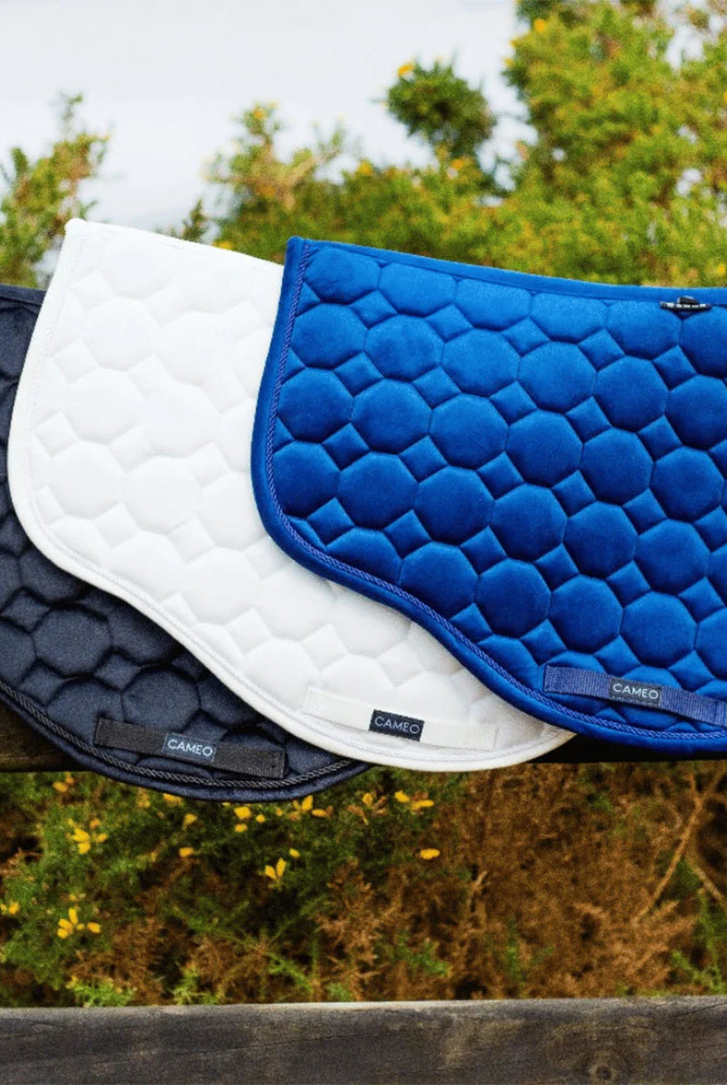 CAMEO Suede GP & Jump Saddlecloth collection lineup in Navy, White, and Black displayed on a wooden fence, showcasing the forward-cut silhouette, geometric octagonal quilting, and rope piping engineered for shock absorption and jump saddle comfort.