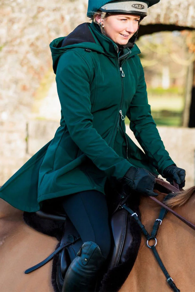CAMEO Zenith Pro Ride Coat in EverGreen modelled ridden on horseback, showcasing the waterproof longline cut, flared rear saddle gusset skirt, high storm collar, and stirrup zips engineered for weather protection and freedom of movement in the saddle.
