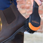 A detailed product photo showing a clean brown leather Chelsea-style yard boot being brushed with a black and orange CAMEO-branded brush. The boot features a subtle embossed CAMEO logo on the side and is paired with a realistic leg wearing navy blue equestrian riding tights.