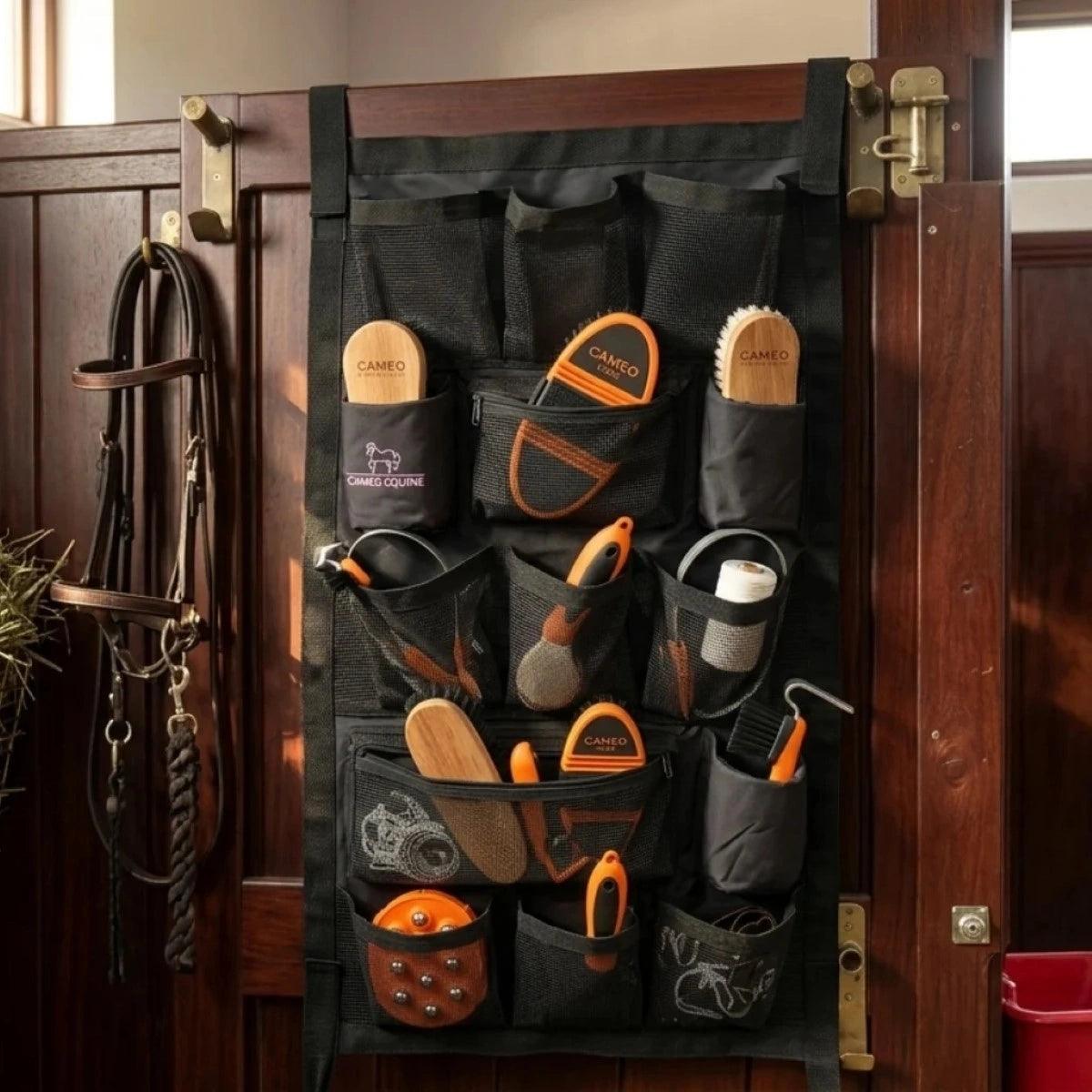 A black equestrian shoe brush organizer hanging on a dark wooden door, holding multiple horse grooming tools including brushes, hoof files, and a leather bridle.