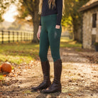 A model wearing dark green equestrian riding breeches with side pockets and a vertical 'CAMEO' logo, paired with dark brown riding boots, standing outdoors on a dirt path with autumn trees and a stone building in the background.