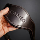 image showing a hand holding the anatomic headpiece in dark brown leather. embossed with 'CAMEO' brand. dark grey background
