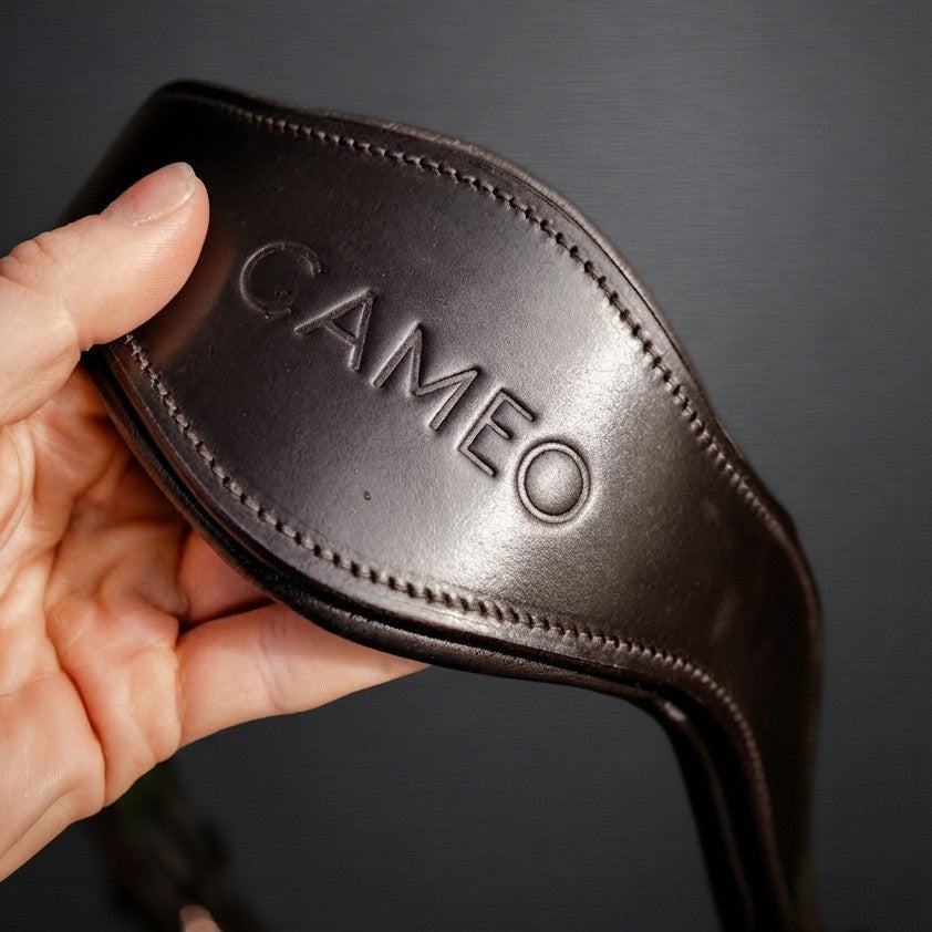 image showing a hand holding the anatomic headpiece in dark brown leather. embossed with 'CAMEO' brand. dark grey background