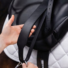 Close-up of a black leather saddle strap being adjusted by hands over a white quilted saddle pad, with the brand name CAMEO embossed on the strap.