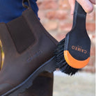 A detailed product photo showing a clean brown leather Chelsea-style yard boot being brushed with a black and orange CAMEO-branded brush. The boot features a subtle embossed CAMEO logo on the side and is paired with a realistic leg wearing navy blue equestrian riding tights.
