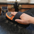 A close-up of a person's hand using an orange and black horse grooming brush on dark horse fur, with a stable interior blurred in the background.
