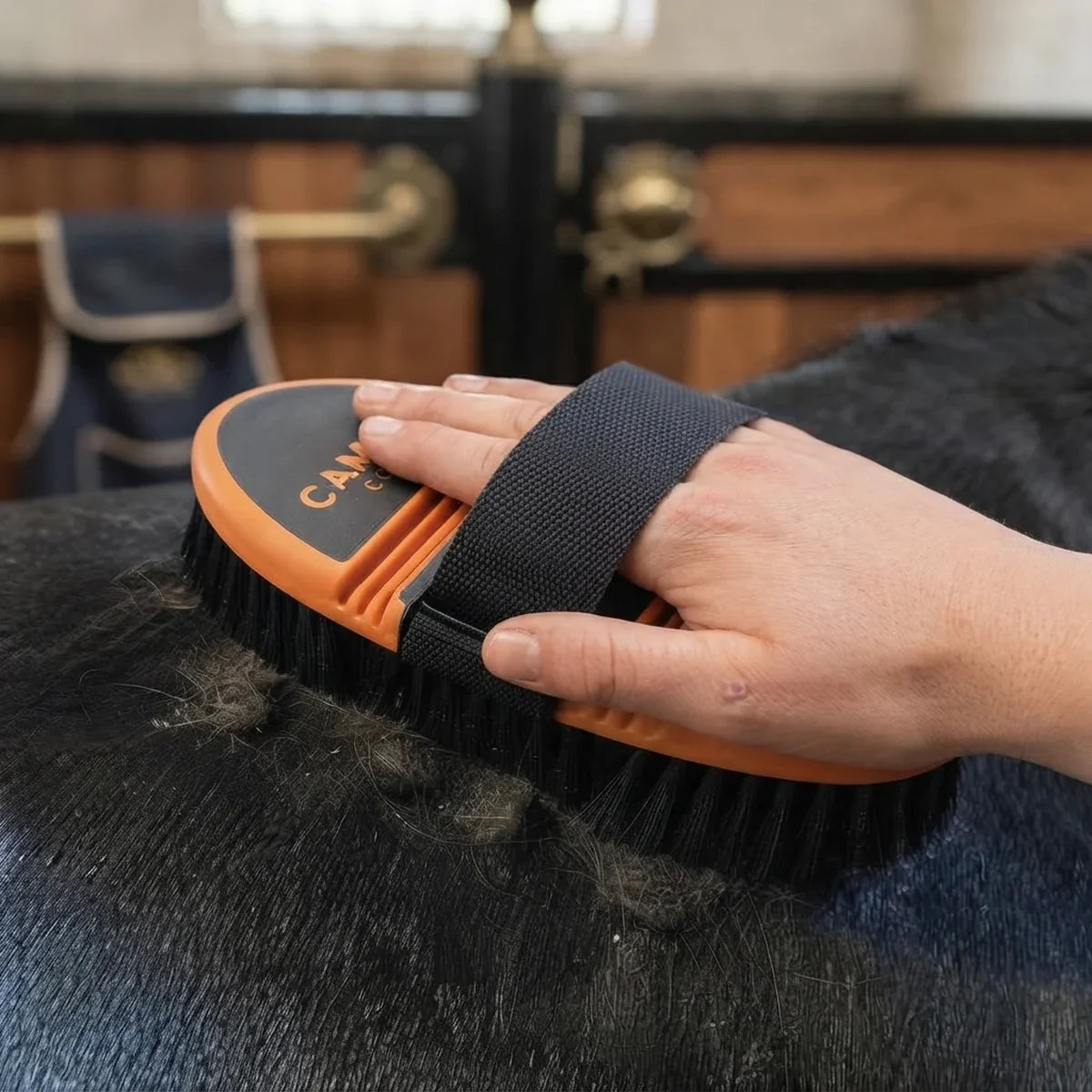 A close-up of a person's hand using an orange and black horse grooming brush on dark horse fur, with a stable interior blurred in the background.