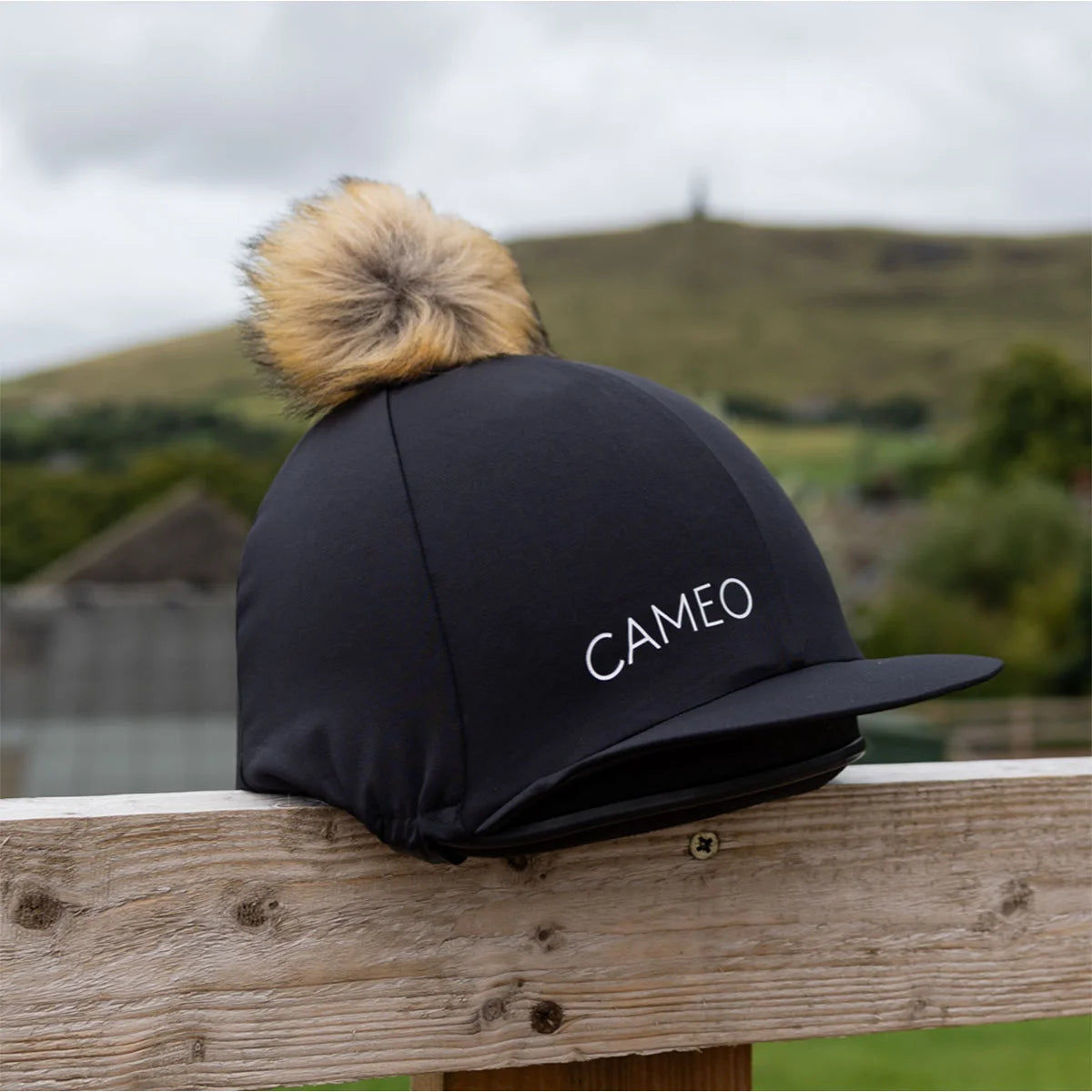 CAMEO Core Hat Silk in Black featuring a luxurious honey-toned faux-fur pom-pom and 4-way stretch technical fabric for a wrinkle-free fit. This premium equestrian helmet cover is designed in the UK with a flexible peak and secure elasticated hem, perfect for matching with the CAMEO CORE collection.