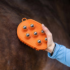 An orange horse grooming brush with six metallic ball nubs being held by a person's hand against a dark brown horse's coat.