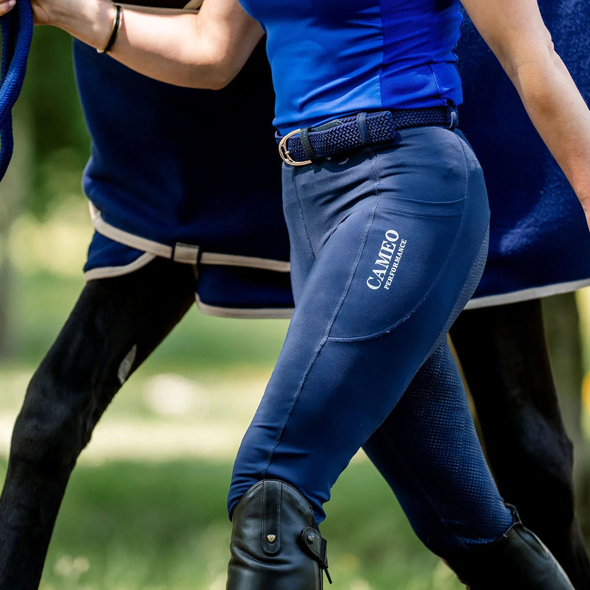 CAMEO Core Woven Stretch Belt in Navy worn with riding tights and tall boots, showcasing the holeless braided elastic weave, leather chapes, and polished stirrup buckle engineered for flexible rider comfort and waistband security in the saddle.