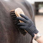 A person wearing a dark gray protective horse grooming glove brushes a dark brown horse's coat with a wooden-handled brush. The brand name CANEO is visible on both the glove and the brush.
