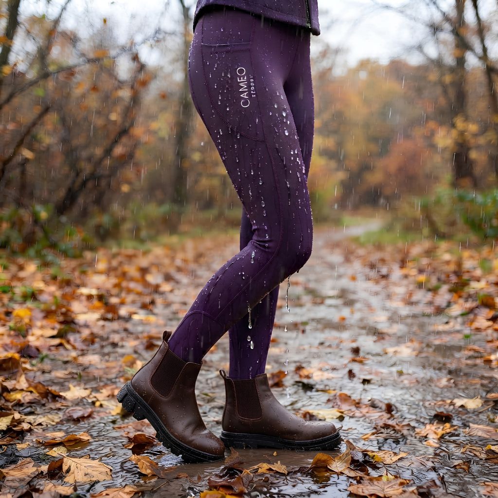 A person wearing purple Caméo leggings and dark brown Chelsea boots stands on a wet leaf-strewn path in the rain, with the brand name visible on the upper thigh.