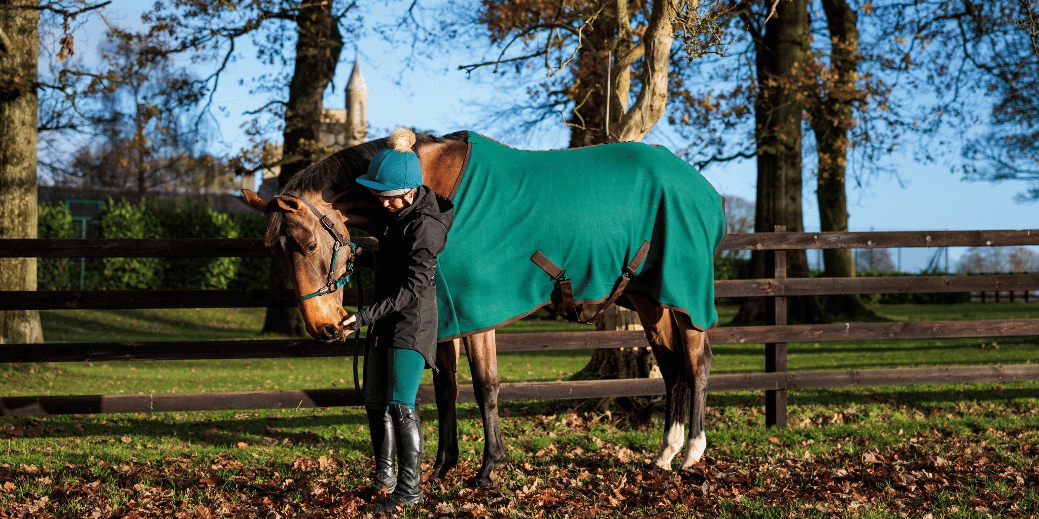A woman wearing a matching green riding hat, jacket, and trousers stands beside a bay horse outdoors, which is wearing a deep green and brown fleece rug from the CAMEO Evergreen AW collection.