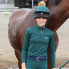 A young rider standing next to a horse, wearing the CAMEO EverGreen C411 kids baselayer, matching green riding bottoms with a dark belt, and a green hat silk with a pompom.