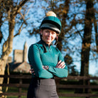 A female equestrian standing outdoors with arms crossed, wearing the CAMEO EverGreen C416 ladies baselayer, dark riding tights with phone pockets, tall leather boots, and a matching teal hat silk with a pompom against a wooded and rustic backdrop.