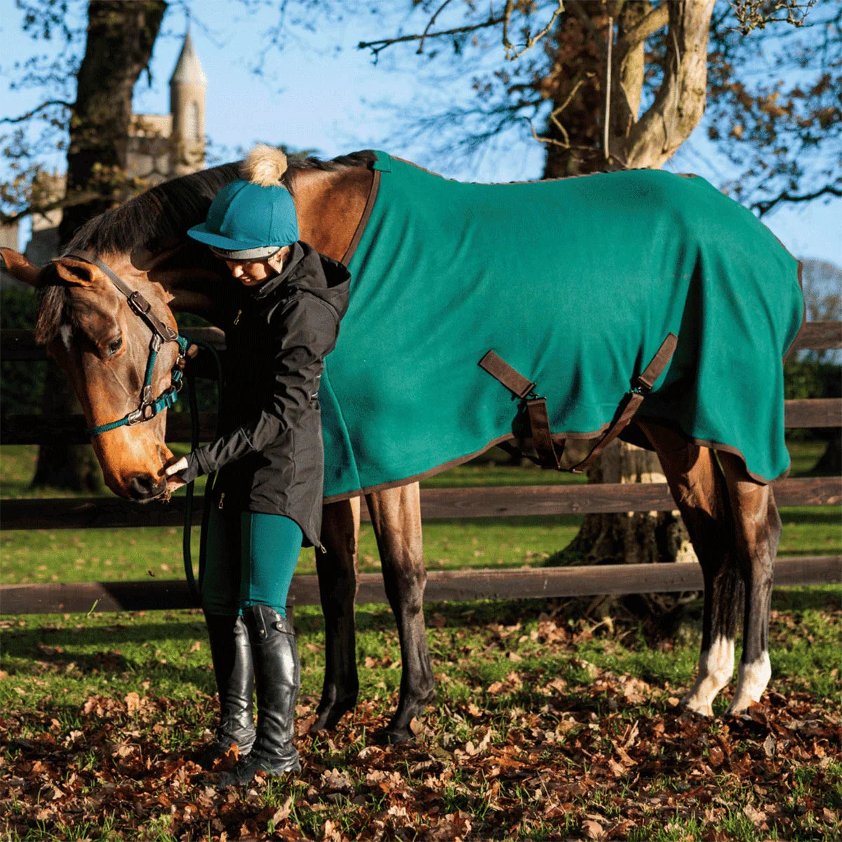 A woman in equestrian attire stands beside a brown horse outdoors, holding the horse's reins while wearing a teal horse blanket with dark brown leather straps. The setting is a grassy field with fallen leaves, wooden fencing, and trees under a blue sky.