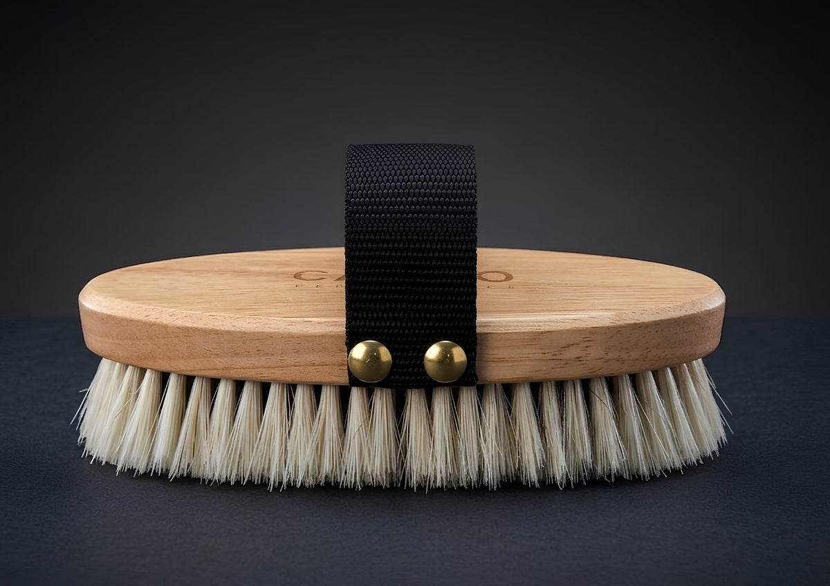 Cameo The Finishing Touch Body Brush featuring a light wood block, dense white bristles, a black webbing hand strap, and brass-finish rivet details on a dark surface.