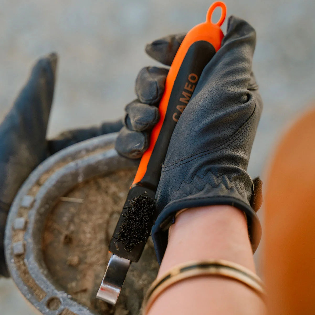 CAMEO Hoof Pick with Brush in Orange in use clearing compacted mud and stones from a shod horse hoof safely.