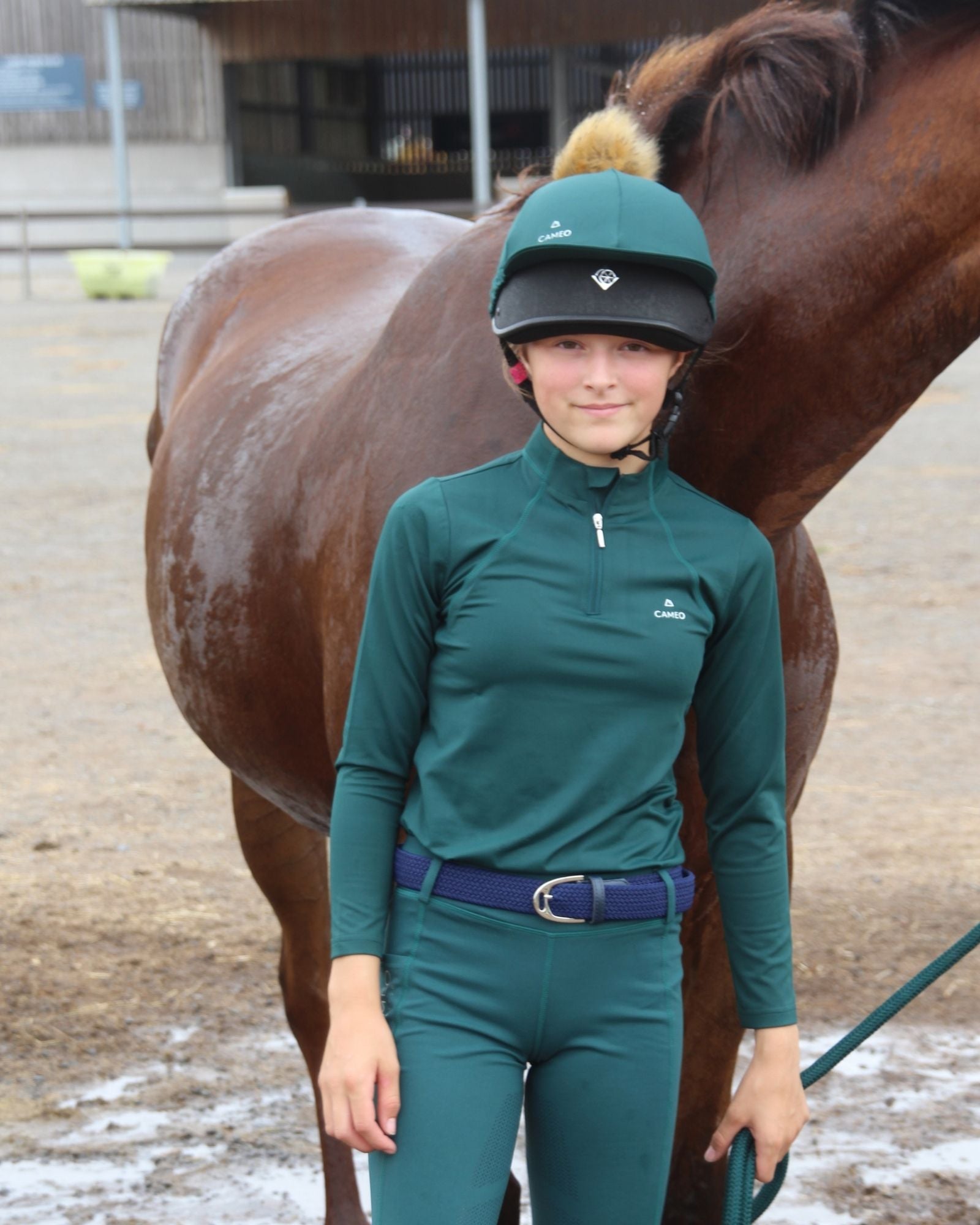 A junior rider wearing a CAMEO deep green technical base layer and matching riding tights, standing next to a horse.