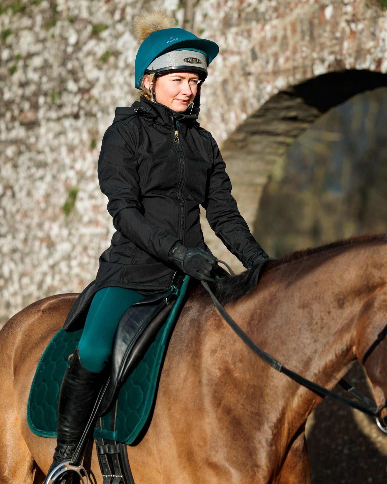 Female equestrian riding a horse wearing the CAMEO black Zenith coat, evergreen All Season riding tights, matching riding hat silk with a faux-fur pompom, and Suede Signature saddlecloth.