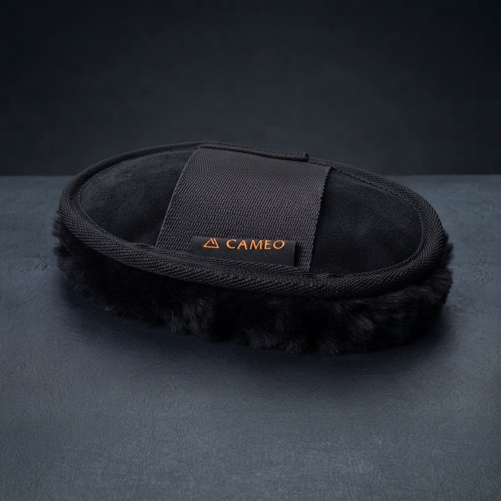 A black lambswool mitt with a black elastic strap featuring the orange 'CAMEO' brand logo, displayed on a dark gray textured surface. for polishing horses coats before competition.