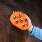An orange horse grooming brush with six shiny silver metal balls, being held by a person's hand against a dark brown horse's coat.