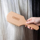 A hand holding a light natural wood paddle hairbrush engraved with 'CAMEO PERFORMANCE' against a background of white and black horse tail hair.