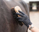 A close-up photograph shows a gloved hand, wearing a dark grey equestrian glove with 'CAMEO' branding on the wrist strap and knuckles, holding a wooden brush against the dark brown coat of a horse. The brush has dark, stiff bristles and is clearly marked with the 'CAMEO' logo on its wooden back. A two-tone watch (gold and silver) is visible on the wrist above the glove. The background is a softly blurred view of a stable aisle