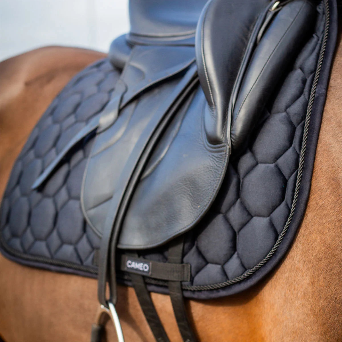 CAMEO Suede Dressage Saddlecloth in Black viewed in an angled close-up beneath a dressage saddle on a horse, highlighting the straight flap cut, honeycomb quilting, rope piping, and reinforced girth loop engineered for shock absorption and non-slip stability during intensive dressage schooling.