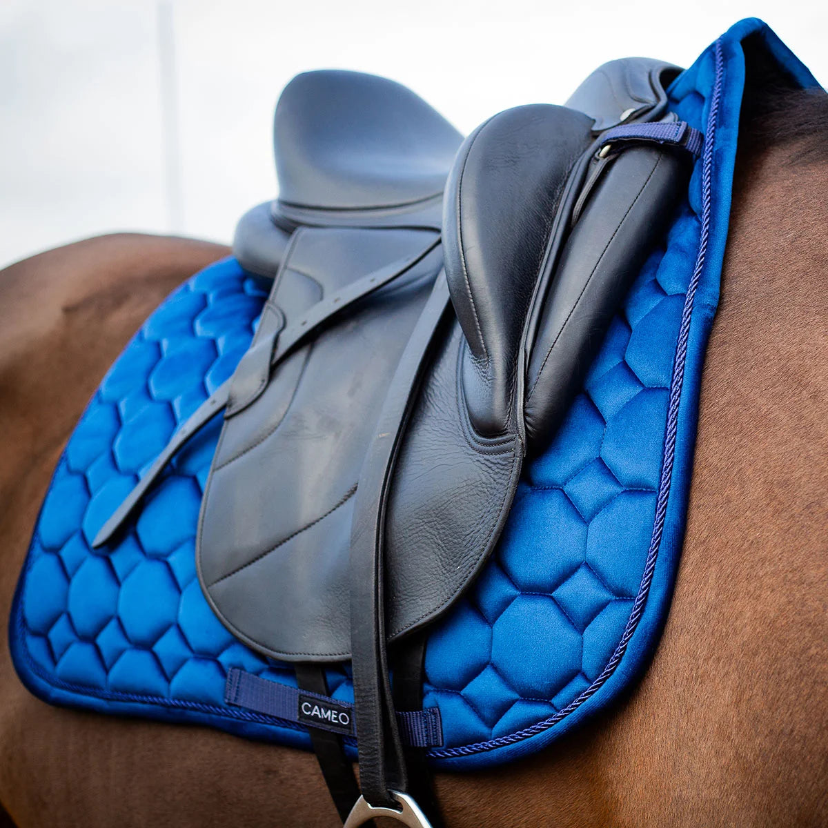 CAMEO Suede Dressage Saddlecloth in Navy viewed in an angled close-up beneath a dressage saddle on a horse, highlighting the straight flap cut, honeycomb quilting, rope piping, and reinforced girth loop engineered for shock absorption and non-slip stability during intensive dressage schooling.