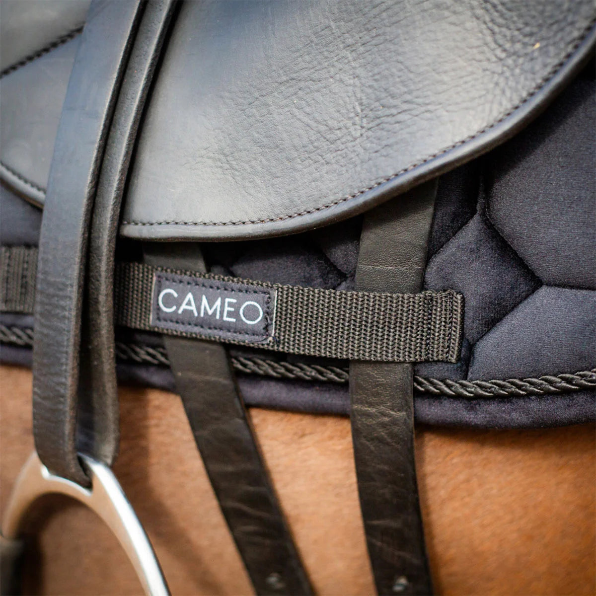 CAMEO Suede Dressage Saddlecloth in Black in a macro close-up beneath a saddle flap, showcasing the heavy-duty webbing girth protection loop with woven brand badge, leather billet straps, and rope piping engineered to prevent saddle friction wear and ensure non-slip stability during dressage riding.