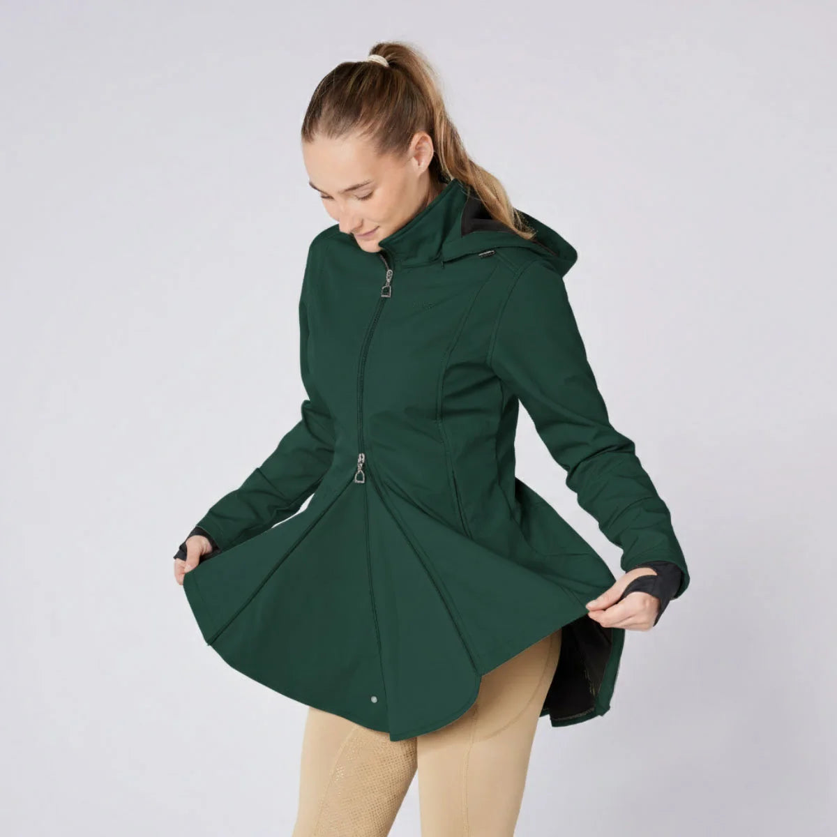 CAMEO Zenith Pro Ride Coat in EverGreen demonstrated by a model holding open the flared riding skirt, showcasing the two-way front zip, thermal fleece lining, and expansive saddle coverage engineered for unrestricted seat and leg aids in wet weather.