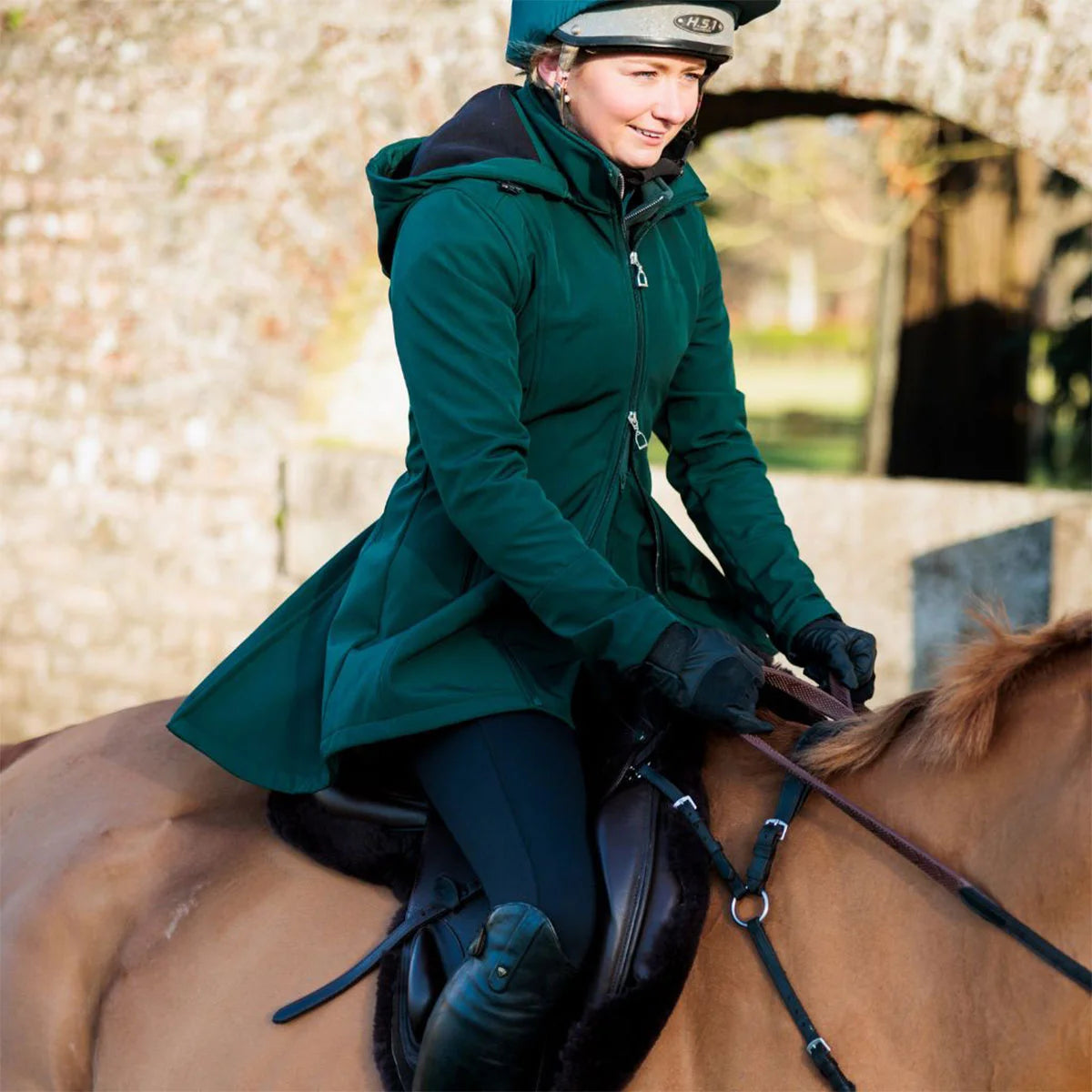 CAMEO Zenith Pro Ride Coat in EverGreen modelled ridden on horseback, showcasing the waterproof longline cut, flared rear saddle gusset skirt, high storm collar, and stirrup zips engineered for weather protection and freedom of movement in the saddle.